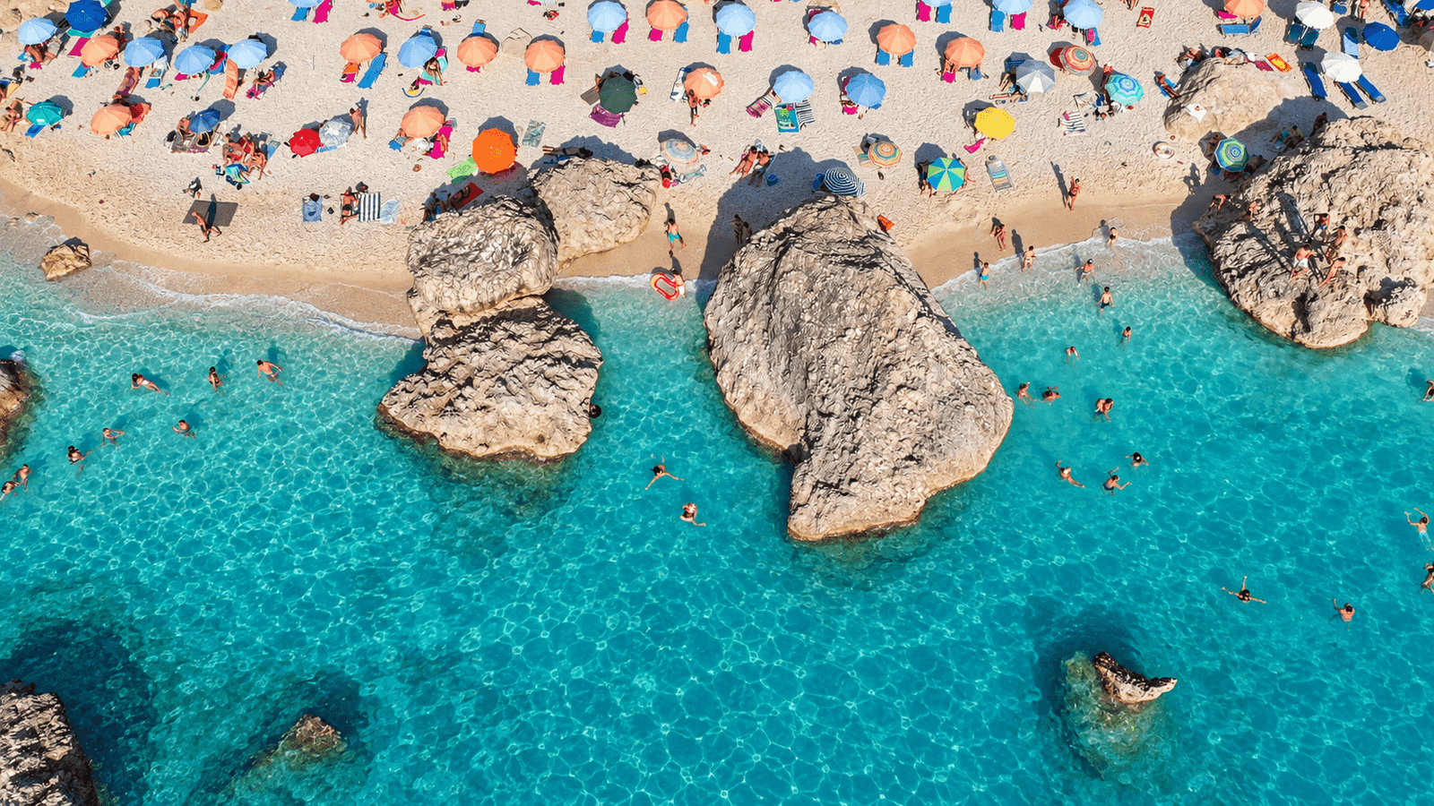 Hidden Beaches in Lefkada 2 Aerial view of large shoreline boulders surrounded by turquoise water, swimmers and colourful beach umbrellas.