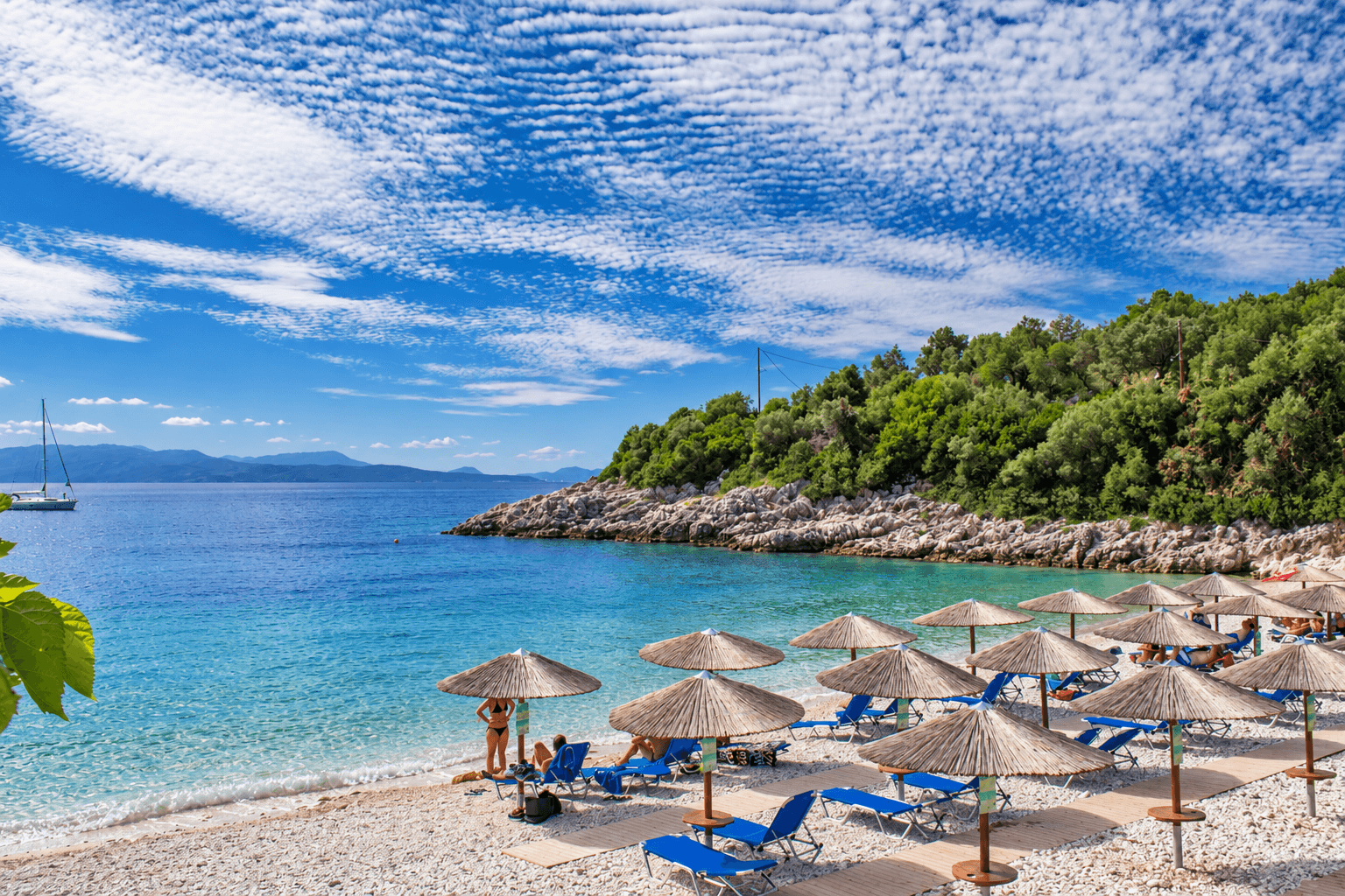 Hidden Beaches in Lefkada 3 Clear turquoise water beside a pebble beach with straw umbrellas, blue loungers and a wooded rocky headland.